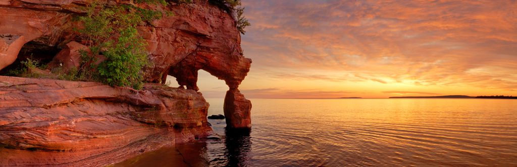 A sandstone arch in Apostle Island National Lakeshore, with a sunset visible in the distance.