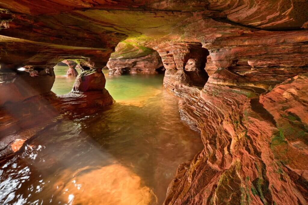 The inside of an Apostle Island National Lakeshore sea cave. It is full of arches, layers of sandstone, and standing water, with rays of sunlight beaming in.