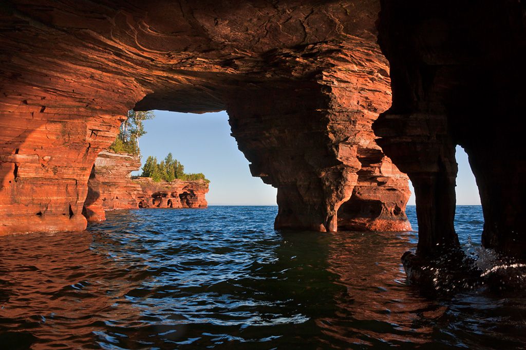 An incredible Apostle Islands photo of a sandstone arch viewed from the water. Devils Islands cliffs are visible in the distance.