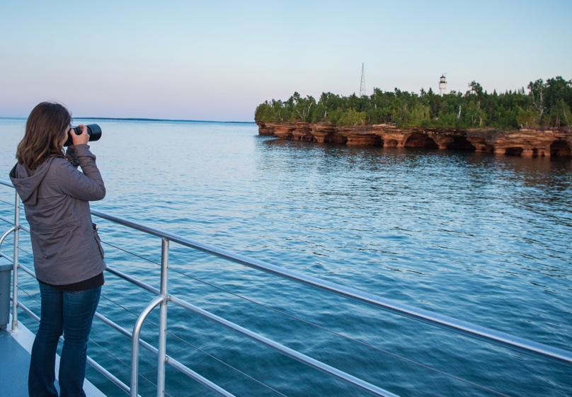 A woman is taking a photo of Devils Island and its lighthouse from the top deck of an Apostle Islands Cruise.