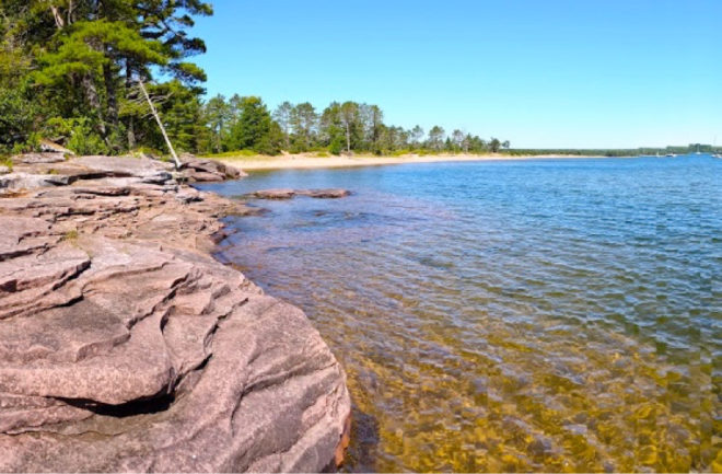 Slate rocky lake shoreline with sandy beach in the background.