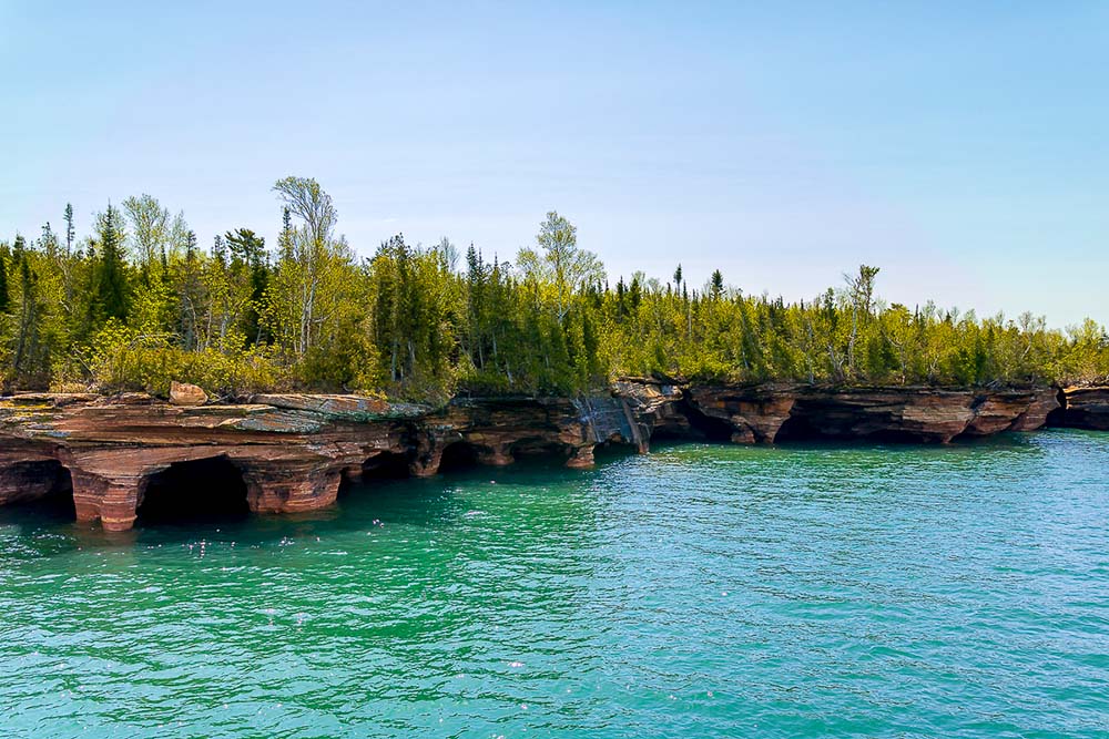 The cliffs and sea caves of Devils Island, featuring red sandstone. A green forest is on top of these cliffs. 