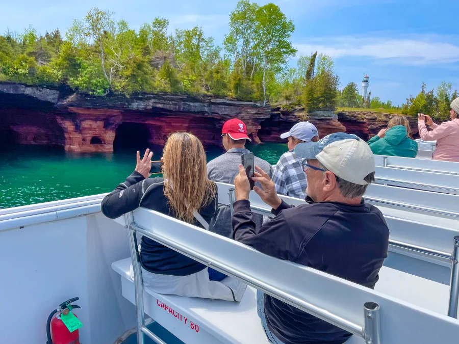 People on the top deck of an Apostle Islands Cruise taking photos of Devils Island, one of Wisconsin's Crown Jewels. The sandstone sea caves are visible and a lighthouse can be seen in the distance, peaking out from trees.